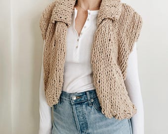 May include: A beige, chunky knit collar scarf. The scarf is worn over a white long-sleeve shirt and blue jeans. The collar scarf is made of a thick, textured yarn and has a wide, folded collar.