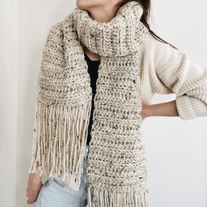 Puede incluir: Bufanda gruesa tejida a mano en hilo beige moteado con flecos. El diseño es de punto grueso. El color neutro permite combinarla fácilmente con diferentes atuendos.