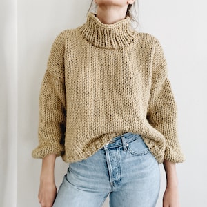 May include: A chunky knit sweater in a light tan color. The sweater features a high turtleneck collar and wide sleeves. The sleeves have a slight ruffle at the cuffs. The sweater is paired with light blue jeans.