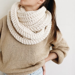 May include: Cream-colored, chunky knit infinity scarf. The scarf is wrapped around the neck and shoulders. The person is wearing a tan sweater and blue jeans. The scarf appears to be hand-knitted with a thick yarn, creating a textured look.
