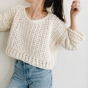 May include: Cream-colored, chunky knit sweater with a wide, rounded neckline and three-quarter sleeves. The sweater has a textured, open weave design and a ribbed hem and cuffs. The sleeves are slightly puffed. The sweater is paired with blue jeans.