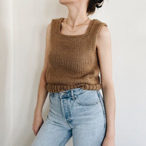 May include: A brown knitted sleeveless top with a square neckline and ribbed detailing. The top has a fitted shape and a ribbed hem. The model is wearing light blue jeans. The top is a neutral colour and can be paired with many outfits.