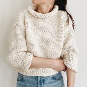 May include: Cream-colored, chunky knit sweater with a wide, rolled turtleneck and long sleeves. The sweater has a relaxed fit and a thick, textured appearance. The sleeves are cuffed at the wrists, and the bottom hem is also thick.