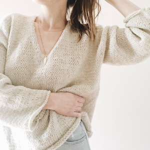 May include: A beige, oversized knit sweater with a deep V-neck and long, flared sleeves. The sweater has a relaxed fit and a textured appearance. A delicate gold necklace with a pendant is visible. The sweater is paired with light-wash denim jeans.