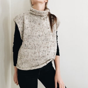 May include: A sleeveless, cream-colored knit sweater with a turtleneck collar. The sweater has a speckled pattern with flecks of brown and other colors. The model is wearing a black long-sleeved shirt underneath and black jeans.