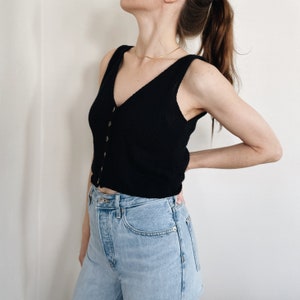 Op de afbeelding: Een zwarte, mouwloze gebreide top met een V-hals en knoopsluiting. De top wordt gecombineerd met lichtblauwe spijkerbroeken. Een goudkleurige ketting is zichtbaar. De outfit wordt gepresenteerd tegen een effen witte achtergrond.