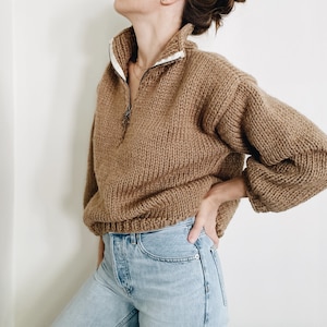 May include: A brown knitted jumper with a half-zip and a white collar. The jumper has long sleeves and a ribbed hem. The zipper is silver. The jumper is paired with light blue jeans.
