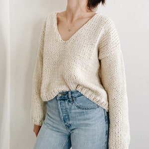 May include: Cream-colored, oversized knit sweater with a deep V-neck. The sweater has long sleeves and a chunky knit texture. The model is wearing light blue jeans. The sweater is a cozy and comfortable garment.