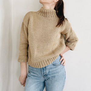 May include: A tan-colored, hand-knitted sweater with a high, ribbed collar and three-quarter sleeves. The sweater has a thick, textured knit and a ribbed hem. The sleeves are cuffed at the elbow. The sweater is paired with blue jeans.