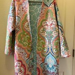 May include: A long, open-front quilted jacket with a vibrant paisley pattern in shades of pink, orange, green, and blue. The jacket is hanging on a black hanger against a neutral background.