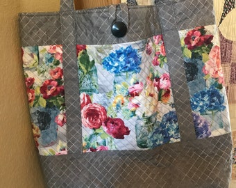 May include: A gray quilted tote bag with floral fabric panels featuring roses and hydrangeas in mason jars. The bag has a button closure and sturdy handles. The overall design is a mix of gray and colorful floral patterns.