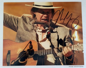 May include: A signed photograph of a musician playing a brown acoustic guitar with a microphone and harmonica. The musician is wearing a light brown jacket, a white shirt, and a light brown cowboy hat. The signature is in black ink.