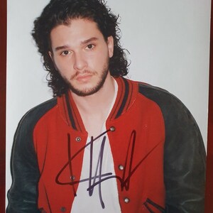 May include: A close-up of a man wearing a red and black varsity jacket. He has dark hair and a beard. The jacket has a white zipper and a white collar. The man is looking at the camera and has a serious expression on his face. The jacket is signed with a purple signature.