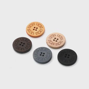 May include: Five round buttons in various colors: light brown, dark brown, gray, and black. Each button has four holes and the engraved text "UGG" repeated around the center. The buttons are made of wood.