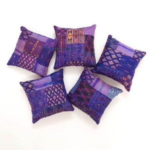Op de afbeelding: Vijf vierkante decoratieve sierkussens in paarse tinten, gerangschikt op een witte ondergrond. Elk kussen heeft een patchwork ontwerp met verschillende patronen en texturen, wat een unieke en artistieke uitstraling creëert. De kussens zijn waarschijnlijk gemaakt van stof.