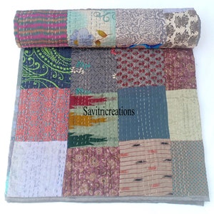 Op de afbeelding: Een patchworkdeken gemaakt van verschillende stoffen in tinten blauw, groen, rood en bruin. De deken is met de hand gestikt en heeft een rustieke, vintage uitstraling. De deken is bovenaan opgerold en de tekst "Savitricreations" is zichtbaar in het midden van de deken.