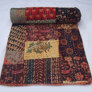 May include: A patchwork quilt featuring squares of patterned fabric in red, brown, and blue hues. The quilt is rolled up at the top, displaying the fabric patterns. It is hand-stitched with white thread, showcasing a variety of designs.