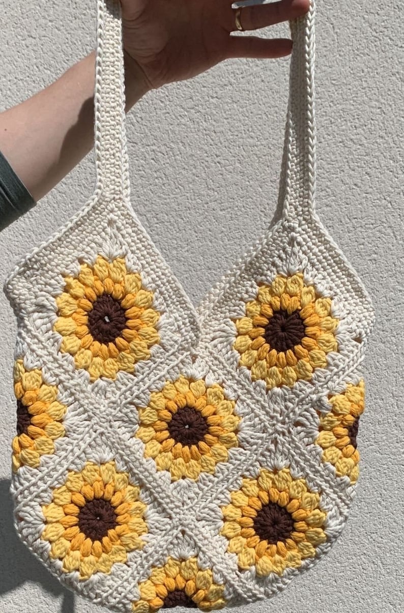 May include: A crocheted tote bag with a sunflower design. The bag is made with white yarn and features yellow and brown sunflowers.