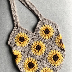 May include: A crocheted tote bag with a sunflower design. The bag is made with beige, yellow, and brown yarn. The bag has a wide, flat bottom and two long straps.