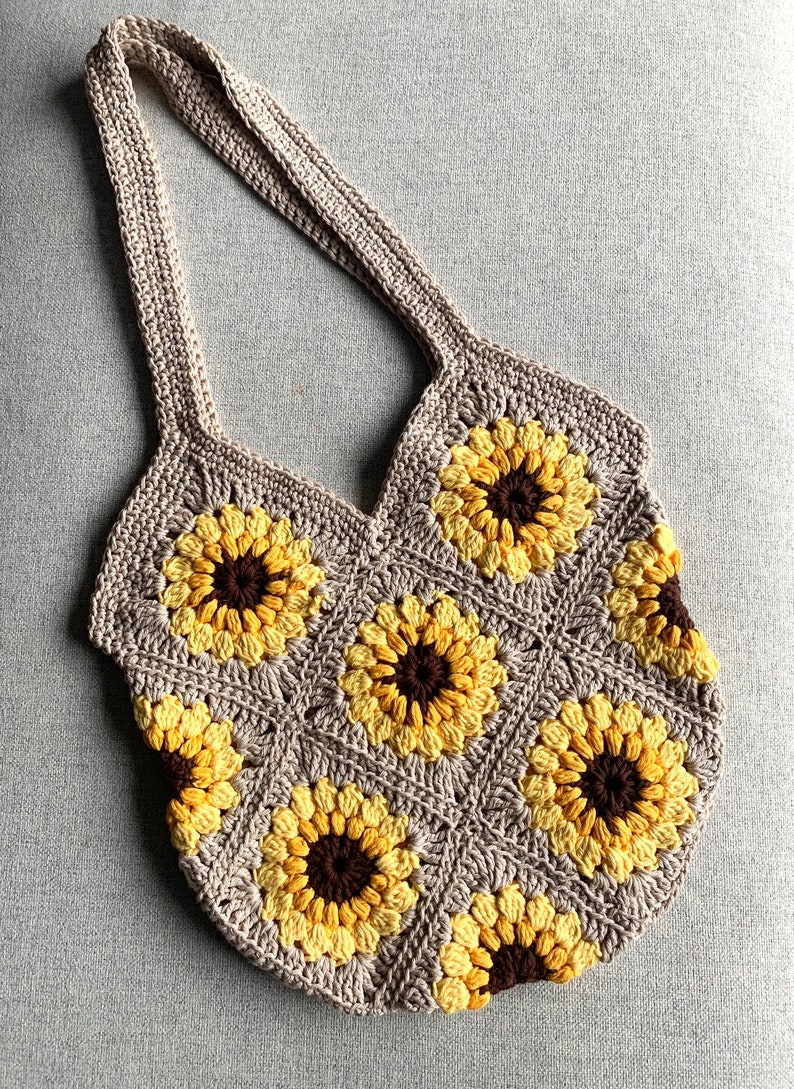 May include: A crocheted tote bag with a sunflower design. The bag is made with beige, yellow, and brown yarn. The bag has a wide, flat bottom and two long straps.