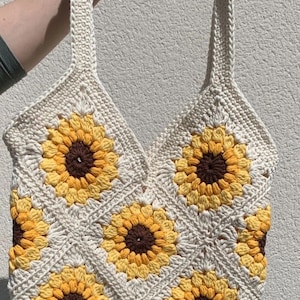 May include: A crocheted tote bag with a sunflower design. The bag is made of cream-colored yarn and features yellow and brown sunflowers.