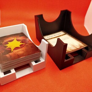 May include: Two 3D printed card holders, one white and one brown. The white holder has a deck of cards inside. The brown holder has a single card inside. Both holders have a curved design to hold the cards.