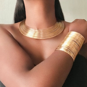 Puede incluir: Un collar choker dorado y una pulsera a juego. El collar es ancho y está hecho de muchas bandas delgadas superpuestas. La pulsera tiene un diseño similar y se lleva en la muñeca.