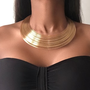 May include: A gold-toned statement necklace with a wide, layered design. The necklace is made of multiple thin metal bands that are stacked on top of each other to create a thick, rounded shape.