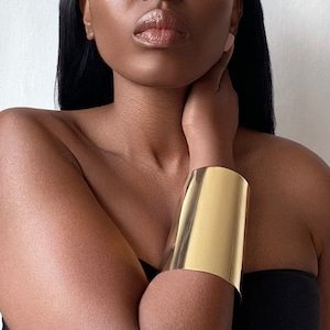 May include: A gold metal cuff bracelet with a smooth, shiny finish. The bracelet is worn on the wrist of a person with dark skin.