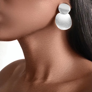 May include: A pair of silver earrings with a round, slightly textured design. The earrings are stacked, with a smaller circle on top of a larger circle.