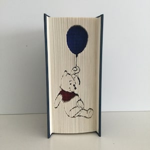 May include: A Winnie the Pooh book folding art piece. The book is folded to create a 3D image of Winnie the Pooh holding a blue balloon. The book is bound in blue.