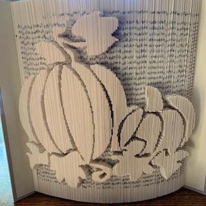 May include: A book sculpture featuring a carved pumpkin design. The pumpkin and leaves are created by folding the pages. The open book reveals the intricate design, ideal for autumn decorations. The pages are white.