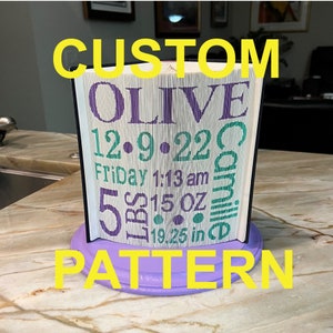 May include: A custom book folding pattern with the name "OLIVE" in purple lettering, the date "12.9.22", the time "Friday 1:13 am", the weight "5 lbs 15 oz", and the length "19.25 in" in green lettering. The book is folded into a 3D shape and is displayed on a purple base.