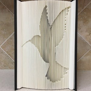 May include: A white book with pages folded to create a 3D hummingbird silhouette. The book is closed and standing upright on a tiled surface.
