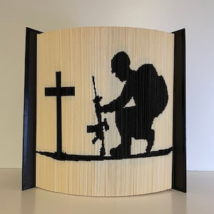 May include: A book sculpture depicting a black silhouette of a soldier kneeling with a rifle and a cross. The artwork is crafted from folded pages, with black borders on each side.