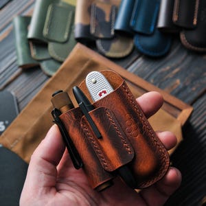 May include: A brown leather multi-tool organizer with a pen, flashlight, and pocket knife. The organizer has a honeycomb pattern and is held in a hand. Other leather organizers in various colors are in the background.