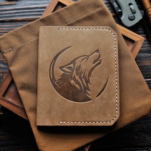 Puede incluir: Cartera de cuero marrón claro con un diseño de lobo aullando grabado en la parte delantera. La cartera rectangular tiene esquinas redondeadas y costuras blancas. Descansa sobre una bolsa de lona marrón y una bandeja de madera.
