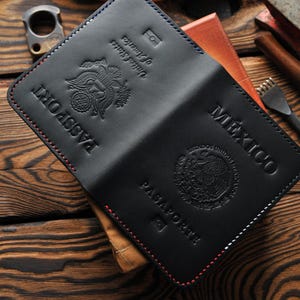 May include: Black leather passport holder with red and blue stitching. The holder is open, displaying the embossed text "PASSPORT" and a United States seal on one side, and "MÉXICO" and a Mexican seal on the other. The holder is on a wooden surface.