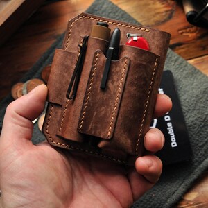 May include: A brown leather wallet with multiple pockets and slots for carrying various items, such as pens, a small knife, and a key chain. The wallet is held in a hand.