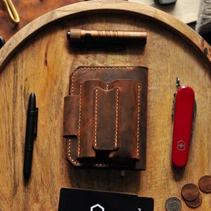 May include: A brown leather wallet with multiple pockets and stitching details. The wallet is on a wooden tray with a red pocket knife, a black pen, a small flashlight, and a business card that says "Double D Leather".