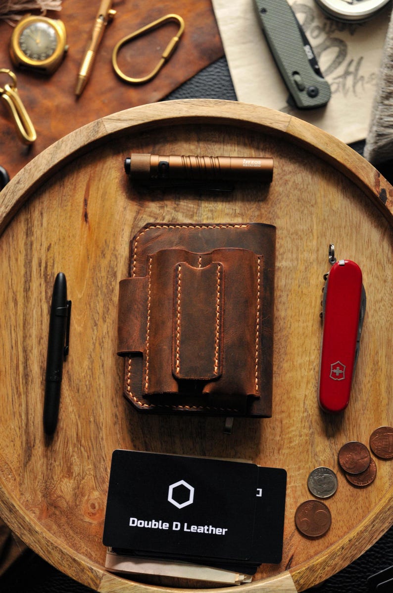 May include: A brown leather wallet with multiple pockets and stitching details. The wallet is on a wooden tray with a red pocket knife, a black pen, a small flashlight, and a business card that says "Double D Leather".