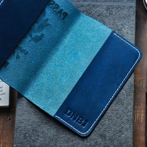May include: A blue leather passport holder with white stitching and the letters "DNBJ" embossed on the front.