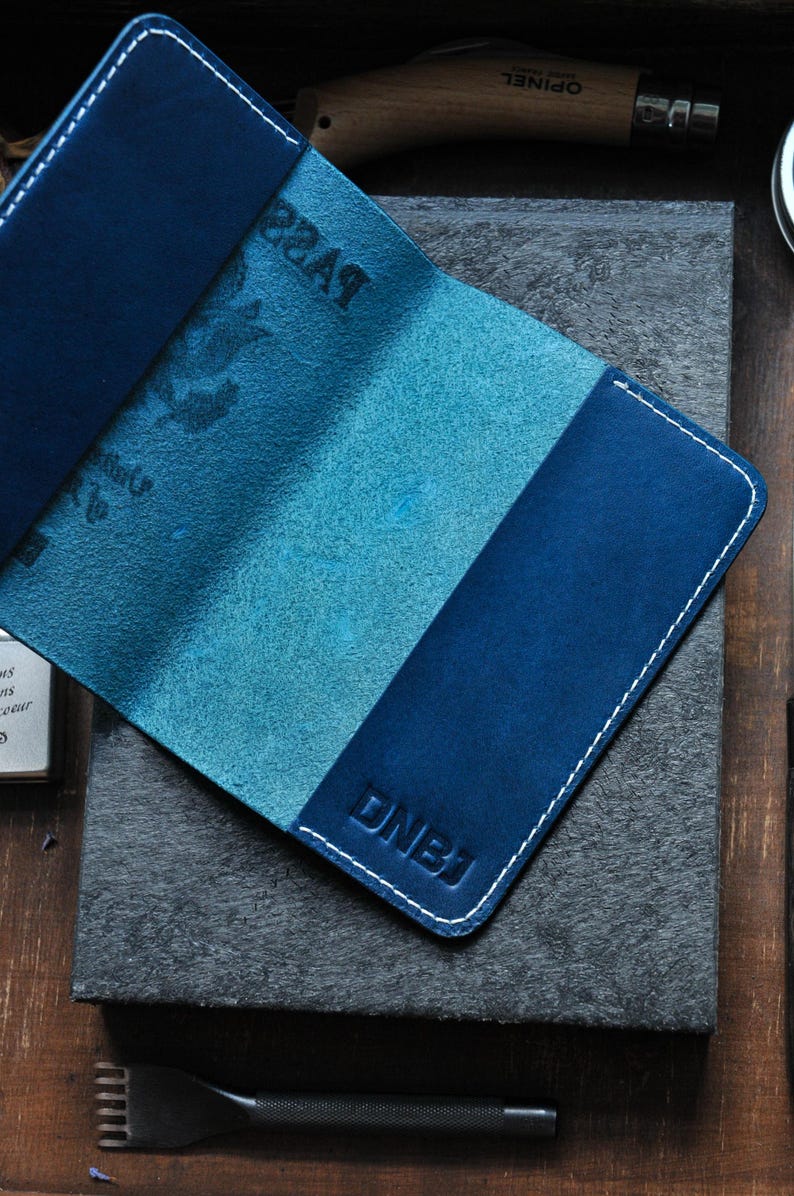 May include: A blue leather passport holder with white stitching and the letters "DNBJ" embossed on the front.