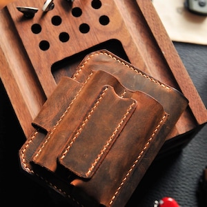 May include: A brown leather wallet with two slots for cards and a flap closure. The wallet is sitting on a wooden surface with a few holes in it.