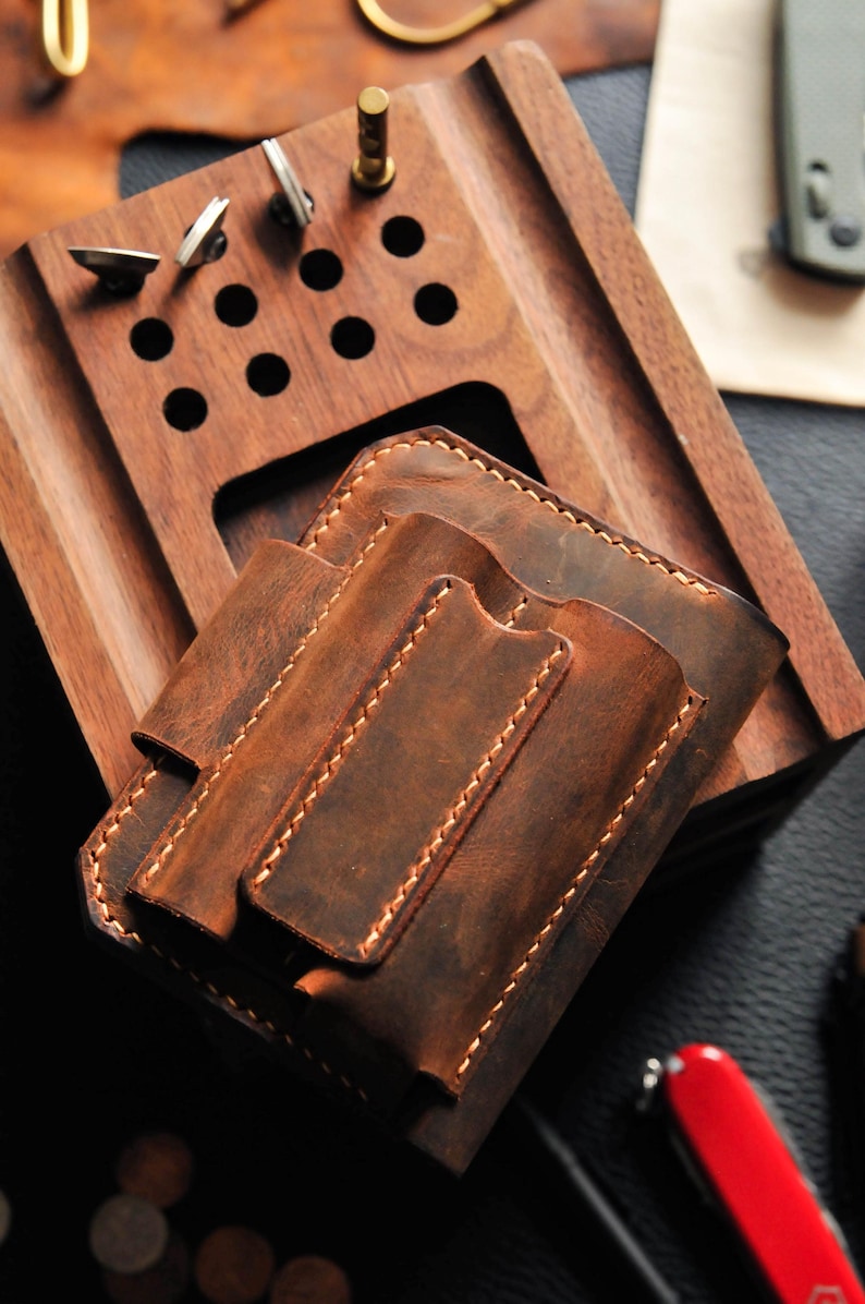 May include: A brown leather wallet with two slots for cards and a flap closure. The wallet is sitting on a wooden surface with a few holes in it.