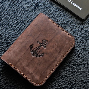 May include: A brown leather wallet featuring an embossed anchor motif. The wallet has a stitched edge and is placed on a textured black surface. A business card with the text "Double D Leather" is also visible.