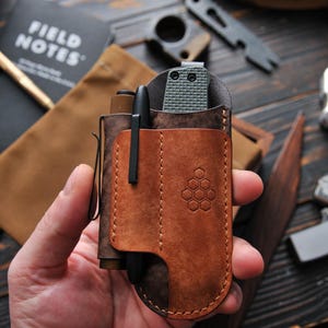 May include: A brown leather pocket organizer with a pen, knife, and other accessories. The organizer has a stitched design and a honeycomb pattern. The Field Notes notebook is visible in the background.