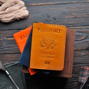 May include: A brown leather passport cover with the words "PASSPORT", "United States of America", and a seal of the United States. The cover is on top of a stack of other passport covers, one orange and one blue.