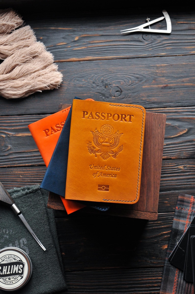 May include: A brown leather passport cover with the words "PASSPORT", "United States of America", and a seal of the United States. The cover is on top of a stack of other passport covers, one orange and one blue.