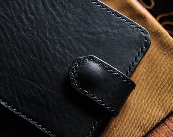 May include: A black leather wallet with a flap closure and stitched edges. The wallet is lying on a brown fabric surface.